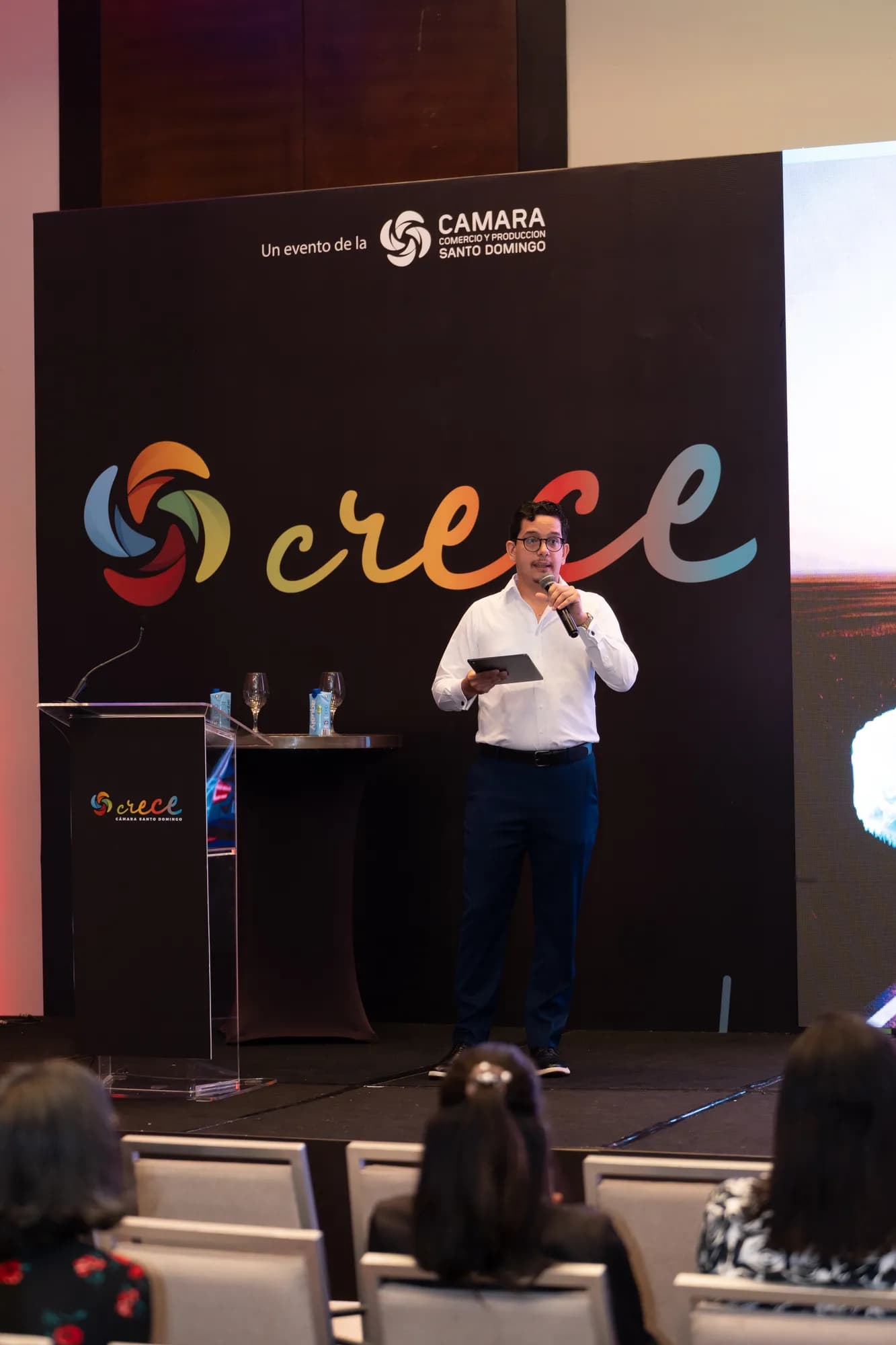 Lucas Pradel on the CRECE 2025 stage holding a tablet and a handheld microphone, in front of the crece backdrop reading 'Un evento de la Cámara Comercio y Producción Santo Domingo'.