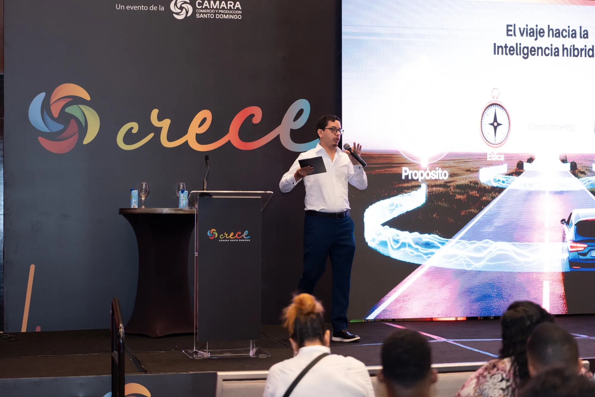 Lucas Pradel speaking on the CRECE 2025 stage beside a screen showing the slide 'El viaje hacia la Inteligencia híbrida', a road illustration labelled Propósito, Ética and Conocimiento.