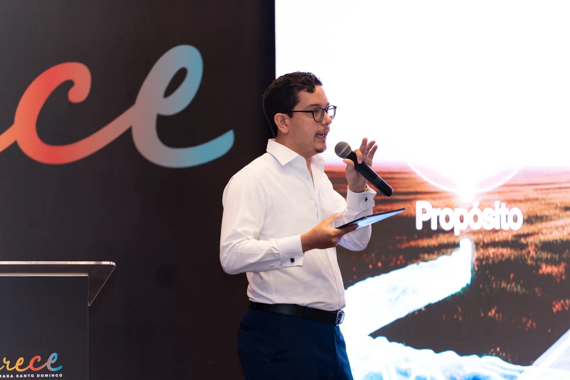 Close view of Lucas Pradel speaking into a handheld microphone at CRECE 2025, with the word 'Propósito' on the screen behind him.