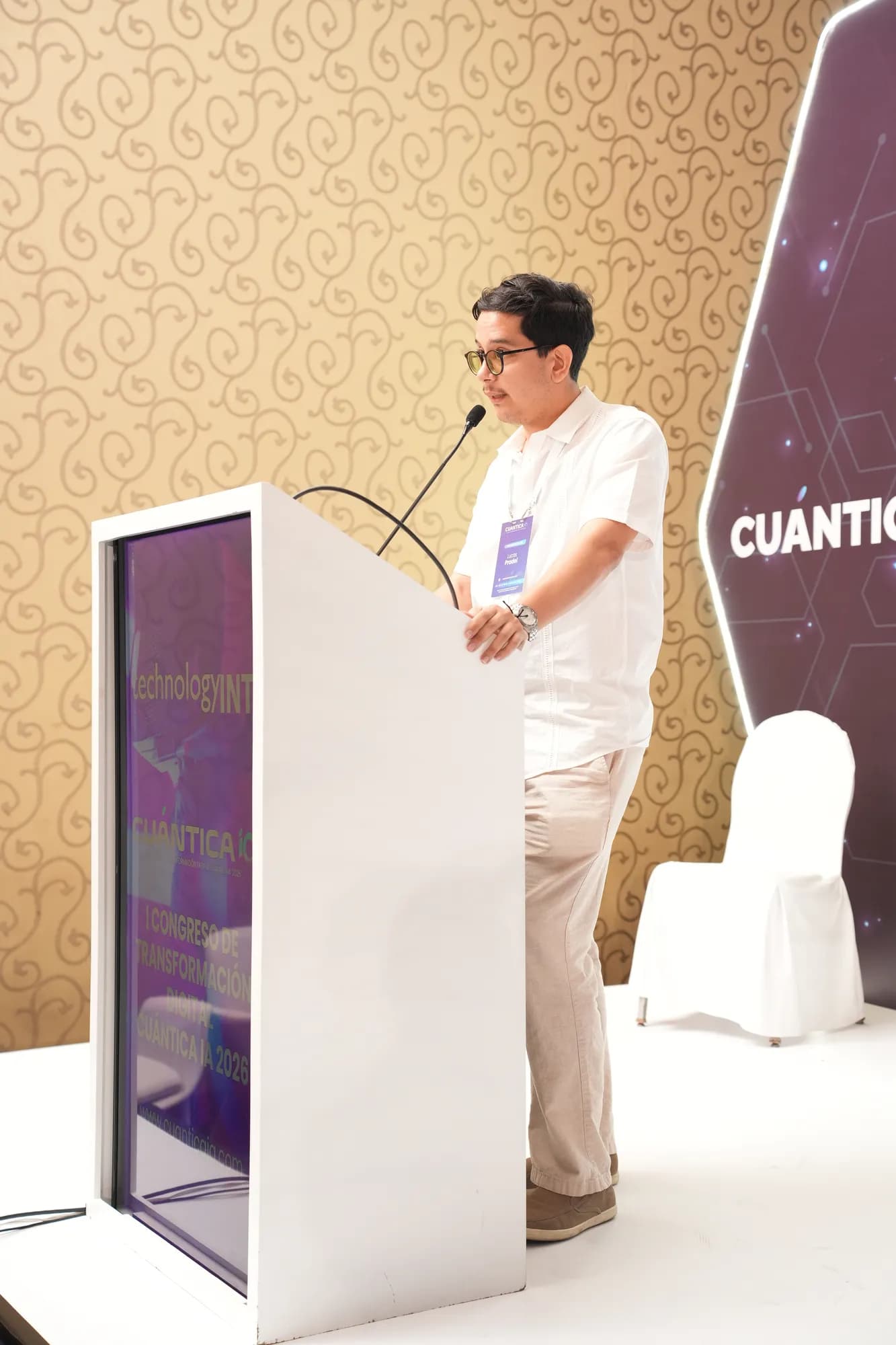 Lucas Pradel mid-talk at the CuánticaIA 2026 podium, wearing a speaker badge and headset microphone.